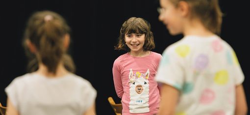 Drei Kinder, zwei davon mit dem Rücken zu uns, unscharf, im Vordergrund, eines zentral, scharf, ein lachendes Mädchen, im Hintergrund sind Stühle zu sehen.