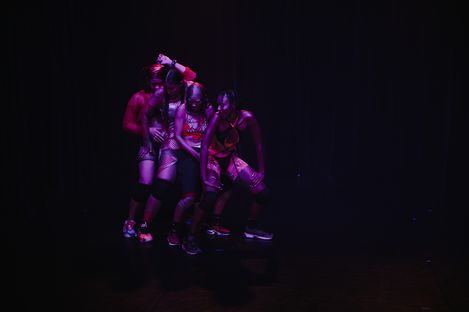 Four people in athletic clothing dancing closely together against a black background with red lighting