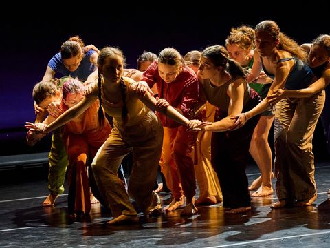 In the foreground is a person with their knees slightly bent and their arms outstretched. Behind them is a group of people touching each other’s arms, their heads bowed.