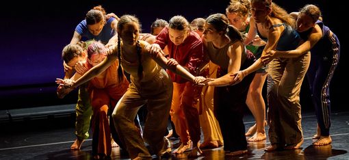 In the foreground is a person with their knees slightly bent and their arms outstretched. Behind them is a group of people touching each other’s arms, their heads bowed.