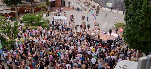 Large crowd in front of an outdoor stage among trees and buildings in daylight