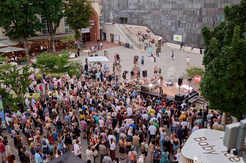 Large crowd in front of an outdoor stage among trees and buildings in daylight