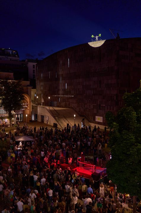 Large crowd in front of a dark, round building with an illuminated person on the roof at night