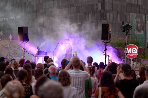 Crowd in front of a stage with fog and purple stage lighting, a building with MQ logo and portrait posters in the background
