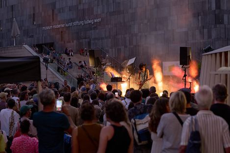 Musician playing guitar and keyboard in front of a large crowd against a modern stone wall with the text 'museum moderner kunst stiftung ludwig wien'
