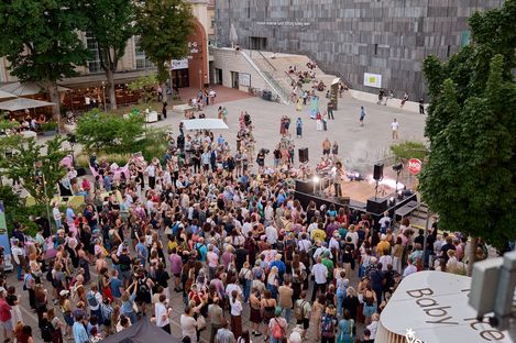 Large crowd in front of an outdoor stage among trees and buildings in daylight