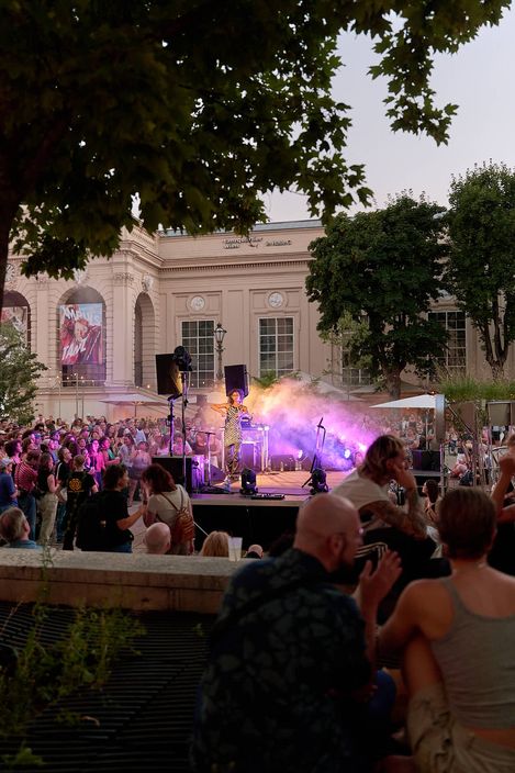 Menschenmenge vor einer Bühne mit Musiker und buntem Bühnenlicht im Freien vor einem historischen Gebäude