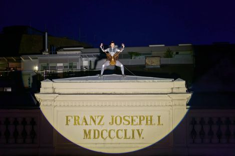 Person in silbernem Anzug mit erhobenen Armen auf einem beleuchteten Gebäudedach mit der Inschrift 'FRANZ JOSEPH I. MDCCCLIV.'
