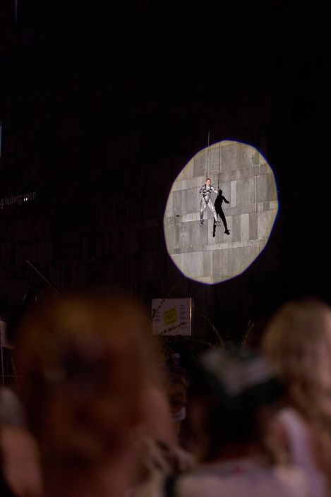 A person in a silver suit is illuminated by a round spotlight on a wall with blurred heads of spectators in the foreground