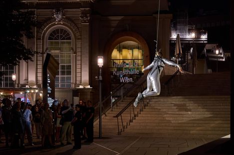 Silver figure in a suit suspended on a rope in front of illuminated stairs with people in the background at night