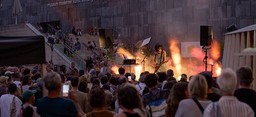 Musikerin mit Gitarre und Keyboard spielen vor einer großen Menschenmenge vor einer modernen Steinwand mit der Aufschrift 'museum moderner kunst stiftung ludwig wien'