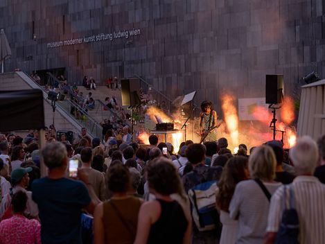 Musikerin mit Gitarre und Keyboard spielen vor einer großen Menschenmenge vor einer modernen Steinwand mit der Aufschrift 'museum moderner kunst stiftung ludwig wien'