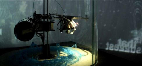 Machine with moving parts and blue net structure on a round platform in a dark room.