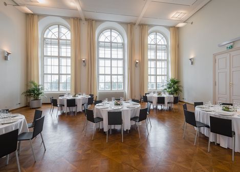 Large bright room with tall arched windows, round tables with white tablecloths and black chairs, parquet floor, and potted green plants