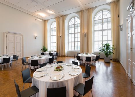 Large bright room with tall arched windows, several round tables with white tablecloths and set places, black chairs, and potted plants