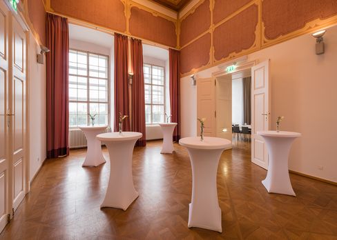 Room in the Baroque Suites at MuseumsQuartier Vienna with tall windows, decorated walls, and cocktail tables covered with white stretch covers each holding a single flower vase.