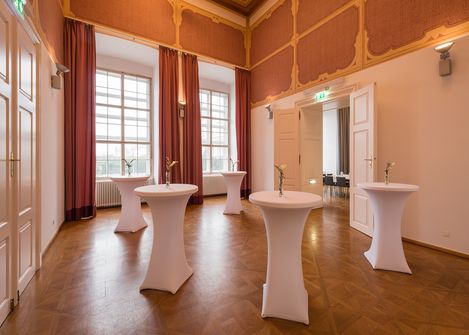 Room in the Baroque Suites at MuseumsQuartier Vienna with tall windows, decorated walls, and cocktail tables covered with white stretch covers each holding a single flower vase.