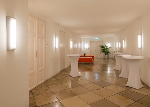 Bright corridor with tall walls, multiple white doors, cocktail tables with white covers, an orange bench, and a large potted plant