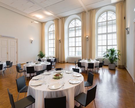 Großer heller Raum mit hohen Rundbogenfenstern, mehreren runden Tischen mit weißen Tischdecken und gedeckten Plätzen, schwarzen Stühlen und Pflanzen in Töpfen