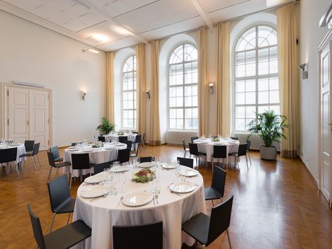 Großer heller Raum mit hohen Rundbogenfenstern, mehreren runden Tischen mit weißen Tischdecken und gedeckten Plätzen, schwarzen Stühlen und Pflanzen in Töpfen