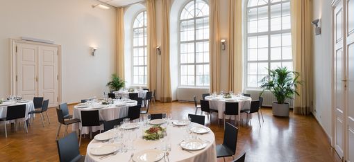 Large bright room with tall arched windows, several round tables with white tablecloths and set places, black chairs, and potted plants