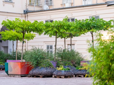 People walking and sitting in front of a historic building with columns and large banners on the facade surrounded by trees and plants in an urban square