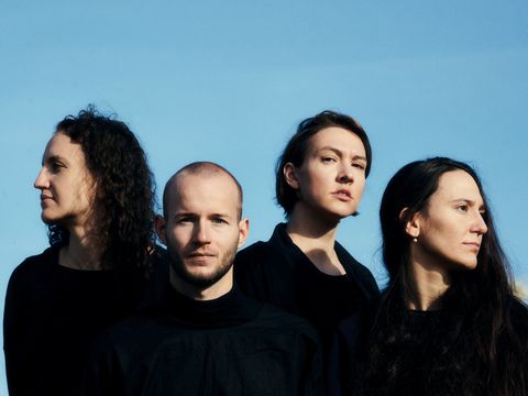 Four people, all dressed in black, are looking in different directions; a blue sky forms the backdrop.