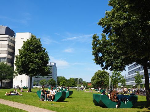Mehrere Personen sitzen und liegen auf grünen Sitzbänken und auf dem Rasen zwischen modernen Gebäuden und Bäumen.