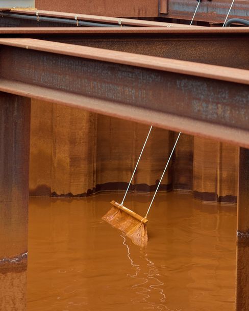 Steel beams above a rust-colored water basin; a wooden board hangs suspended by ropes in the water.