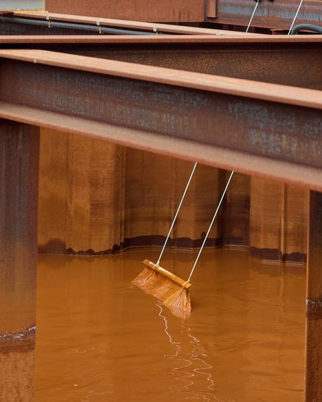 Steel beams above a rust-colored water basin; a wooden board hangs suspended by ropes in the water.