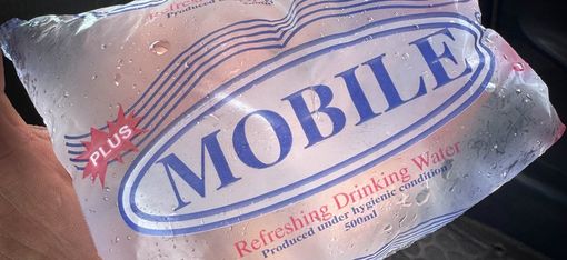 Hand hält eine durchsichtige, mit Wasser gefüllte Plastikverpackung mit der Aufschrift 'MOBILE' und 'Refreshing Drinking Water'
