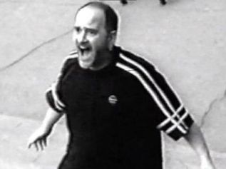 Man with short hair and beard shouting with open mouth and outstretched arms on a stree.