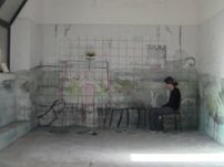 Person sitting at a table in a large empty room with peeling paint and exposed pipes on the wall.