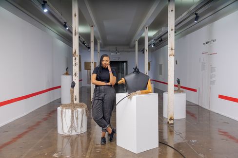 Person leaning against a white pillar in a white exhibition room with a red stripe next to a black dress form and an object with roots on a pedestal.