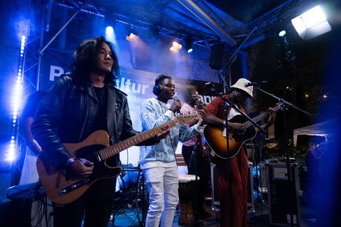 Three musicians on a stage at a concert, one playing electric guitar, one singing into a microphone, one playing acoustic guitar and wearing a hat.