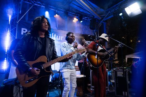 Three musicians on a stage at a concert, one playing electric guitar, one singing into a microphone, one playing acoustic guitar and wearing a hat.