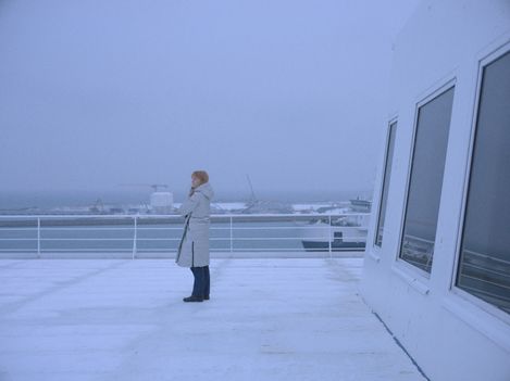 Eine Person steht steht auf einen Schiffsdeck und blickt in die Ferne. Es ist stürmisch und wirkt sehr kalt.