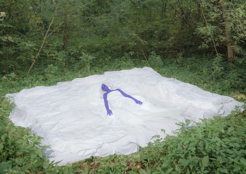 Sculpture of a blue humanoid figure lying on a large irregular white surface in a wooded area.