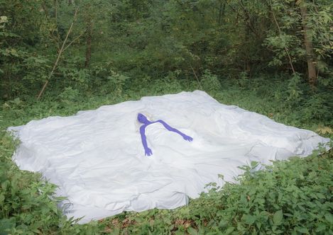 Sculpture of a blue humanoid figure lying on a large irregular white surface in a wooded area.
