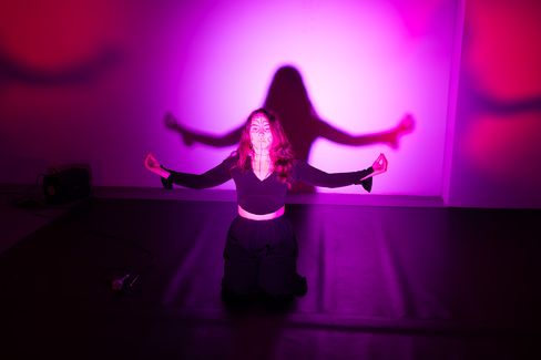 A woman kneels with outstretched arms in front of a pink-purple wall with her shadow clearly visible.