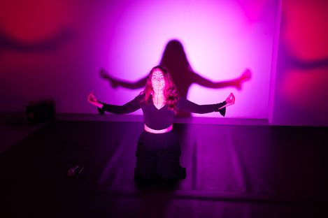 A woman kneels with outstretched arms in front of a pink-purple wall with her shadow clearly visible.
