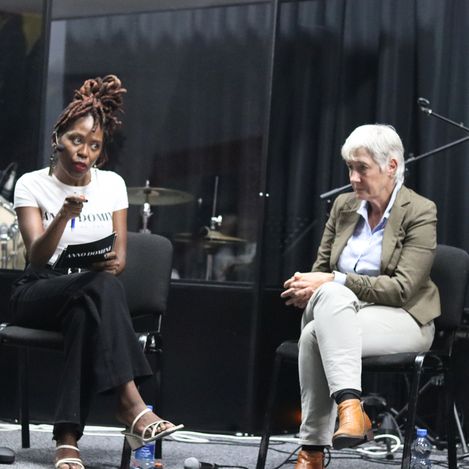 Two people sit on chairs on a stage with a black background, one holding a microphone and a sheet of paper, the other sitting with hands clasped.