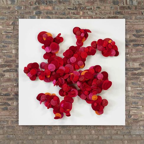Wall installation of many round red and orange felt discs on a white square background against a brick wall.