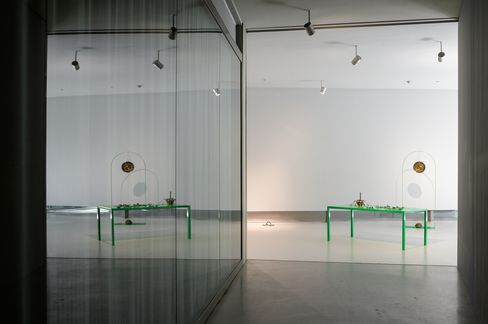 Leerer Ausstellungsraum mit grünem Tisch und Kunstobjekt vor weißer Wand, Spiegelung an Glaswand links.