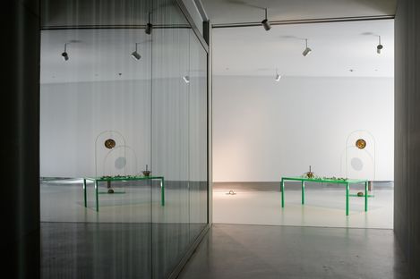 Leerer Ausstellungsraum mit grünem Tisch und Kunstobjekt vor weißer Wand, Spiegelung an Glaswand links.