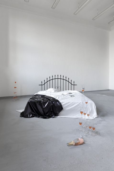 Bed with black and white covers, tipped champagne bottle and several filled champagne glasses on the floor in a white room with high ceiling.