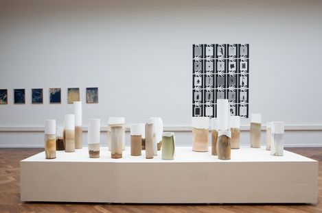 Several cylindrical ceramic objects of various sizes and colors on a white pedestal in an exhibition room with additional artworks on the walls.