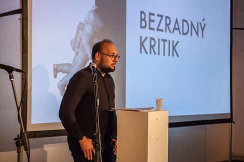 Man speaking at a podium with a microphone in front of a projected slide titled “Bezradný kritik.”