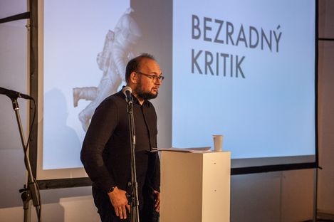 Man speaking at a podium with a microphone in front of a projected slide titled “Bezradný kritik.”