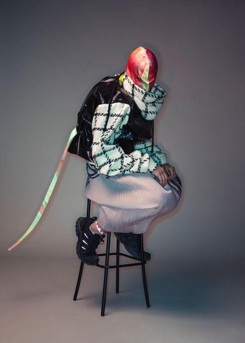 Person wearing a red-and-white patterned headscarf, checkered sweater, black vest, pleated skirt, and black shoes sitting on a stool leaning forward.